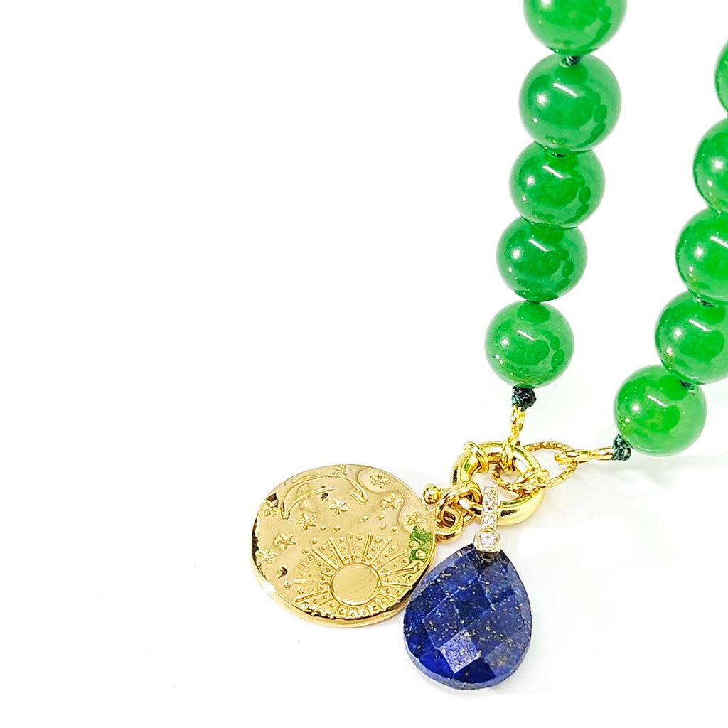 A close-up of a beaded necklace with glossy green beads, a gold-toned engraved disc charm, and a faceted blue lapis lazuli tear-drop pendant on a white background.