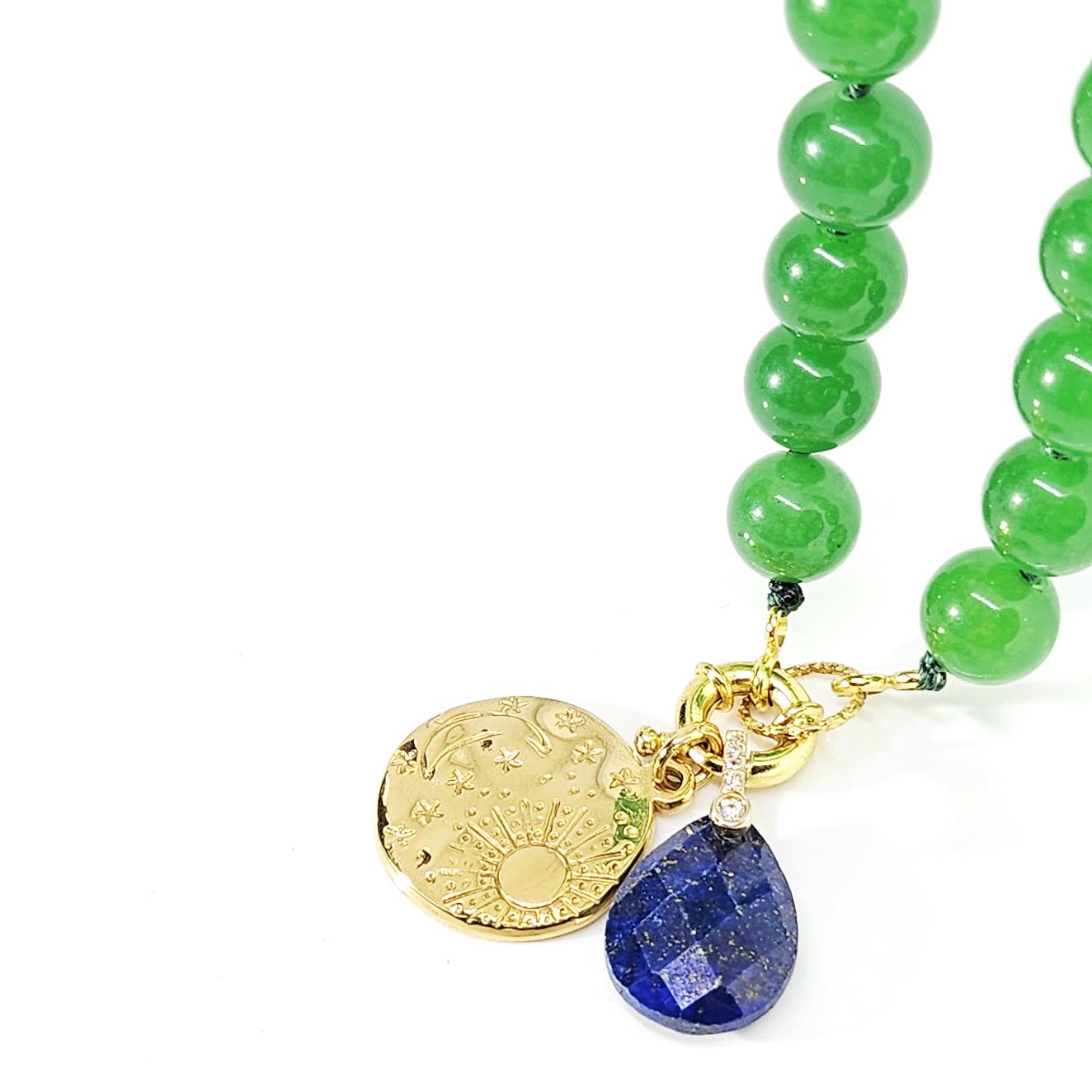 A close-up of a beaded necklace with glossy green beads, a gold-toned engraved disc charm, and a faceted blue lapis lazuli tear-drop pendant on a white background.