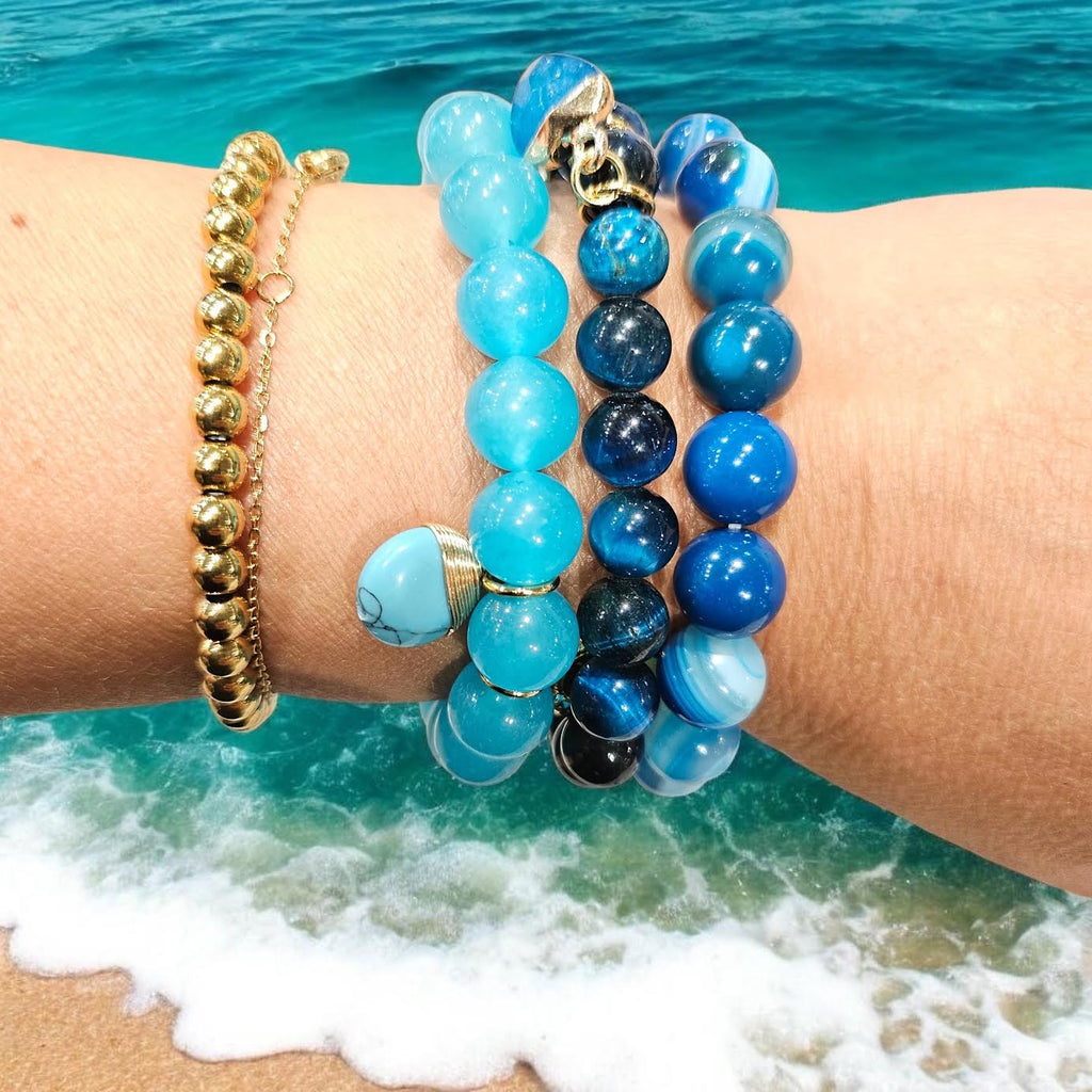 A close-up of a person's wrist wearing multiple beaded bracelets, including blue and turquoise stone bracelets, a gold ball bracelet, and a skull charm, with ocean waves and turquoise water in the background.