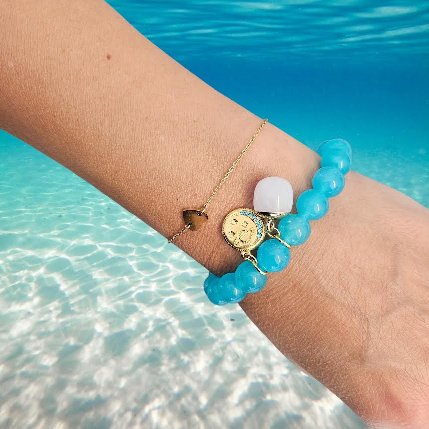A close-up of a person's wrist wearing three bracelets against a tropical ocean background. The bracelets include a beaded turquoise bracelet with a gold moon charm, a gold chain bracelet with a small brown heart, and a gold chain bracelet with a white oval stone.