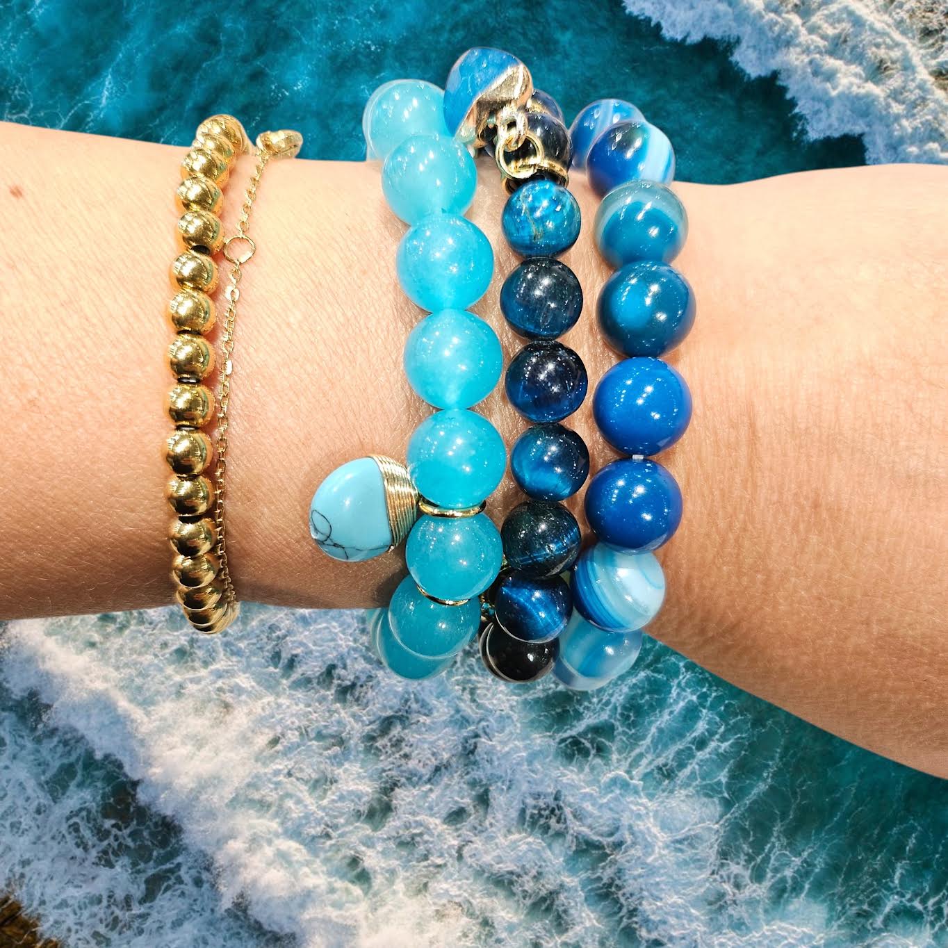 A close-up of a person's wrist wearing multiple beaded bracelets, including blue and turquoise round beads, a skull charm, and a gold ball chain bracelet, with ocean waves in the background.