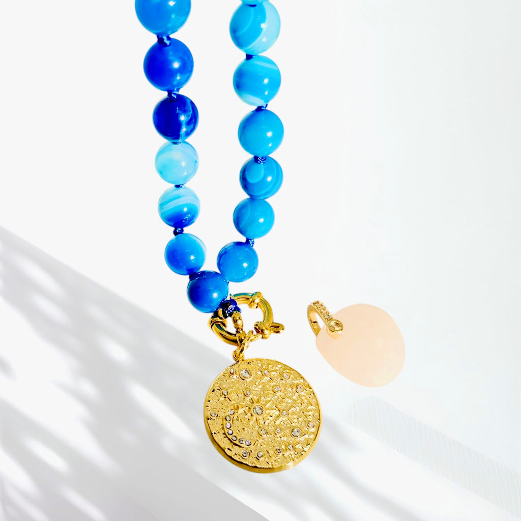 A beaded necklace with light and medium blue beads, a textured gold round pendant, and a pale peach stone charm, photographed on a white background with soft shadows.
