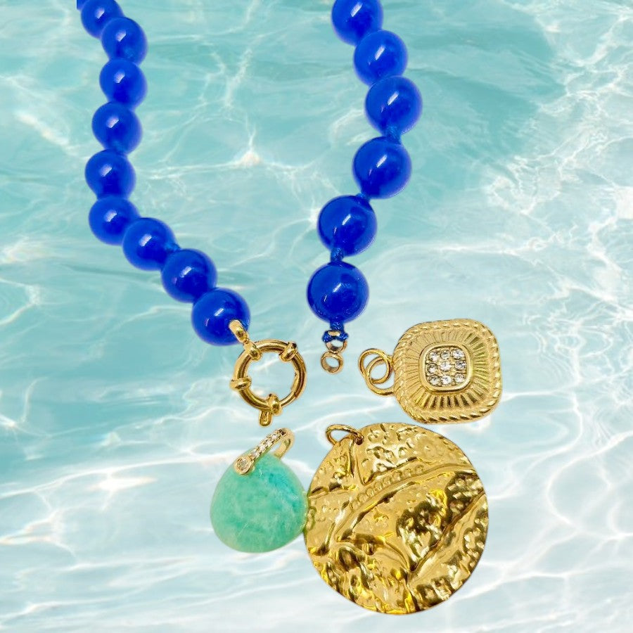 A beaded necklace with glossy cobalt blue beads, gold-tone hardware, and three charms: a round textured gold pendant, a square gold locket-style charm with a clear stone, and a smooth light green teardrop charm, displayed against a turquoise water background.