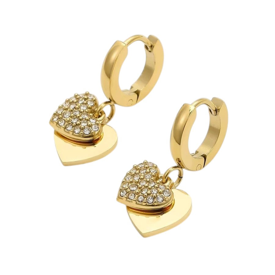 A pair of gold huggie hoop earrings with dangling heart charms. One heart charm is smooth gold, while the other is encrusted with small clear pavé stones, set against a plain white background.
