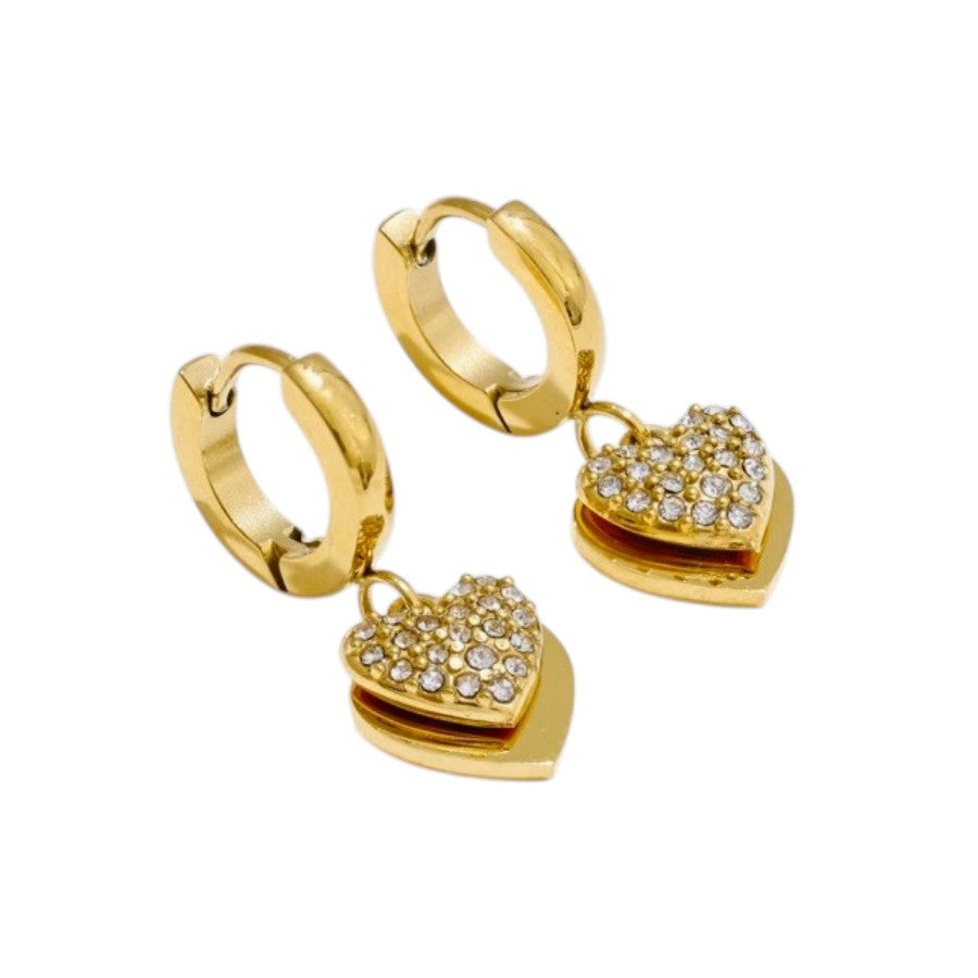 A pair of gold huggie hoop earrings with dangling heart charms. The heart charms are outlined in gold and paved with small clear crystals, with a polished gold center layer visible behind them, set against a plain white background.