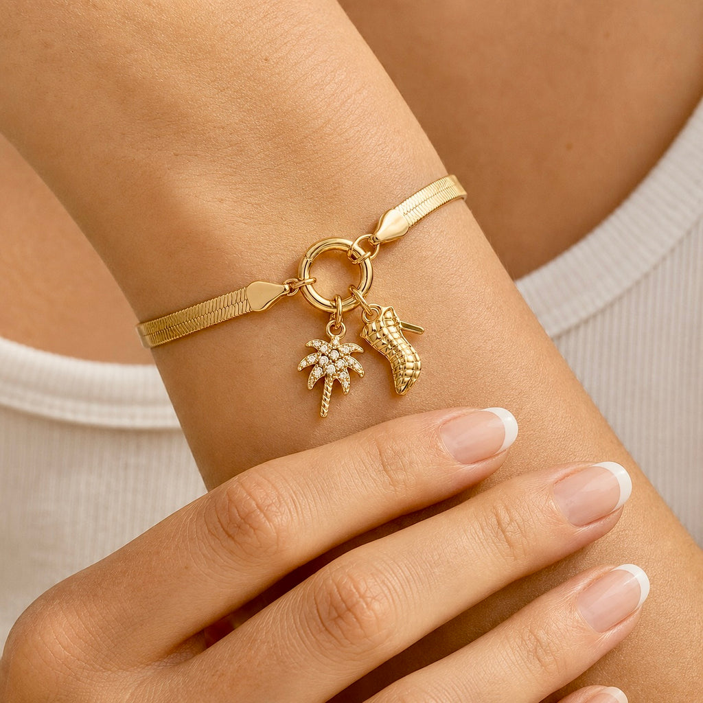 A close-up of a gold bracelet worn on a person's wrist, featuring a textured band, a ring clasp, and three charms: a small palm tree, a coconut shell, and a small flower cluster. The person is wearing a white ribbed top.