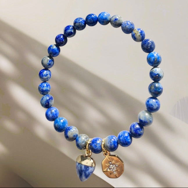 Two beaded stretch bracelets displayed against a light neutral background. The top bracelet features multicolor tiger's eye beads with gold-toned charms, and the bottom bracelet features blue and white marbled beads with gold-toned charms.
