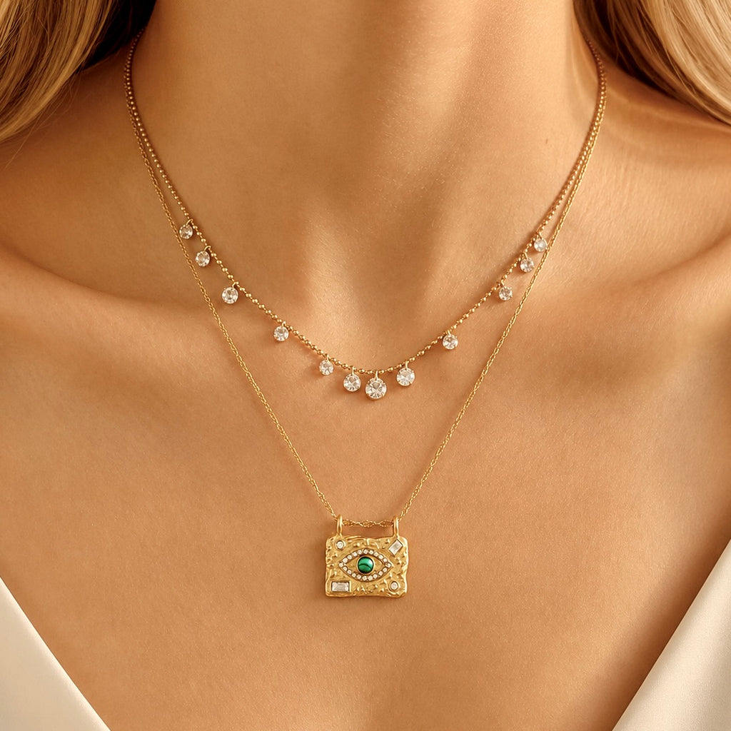 A close-up of a model wearing a delicate gold double-layer necklace. The top layer features small clear crystal drops, while the bottom layer has a textured gold rectangular pendant with a green evil eye design surrounded by tiny clear crystals.
