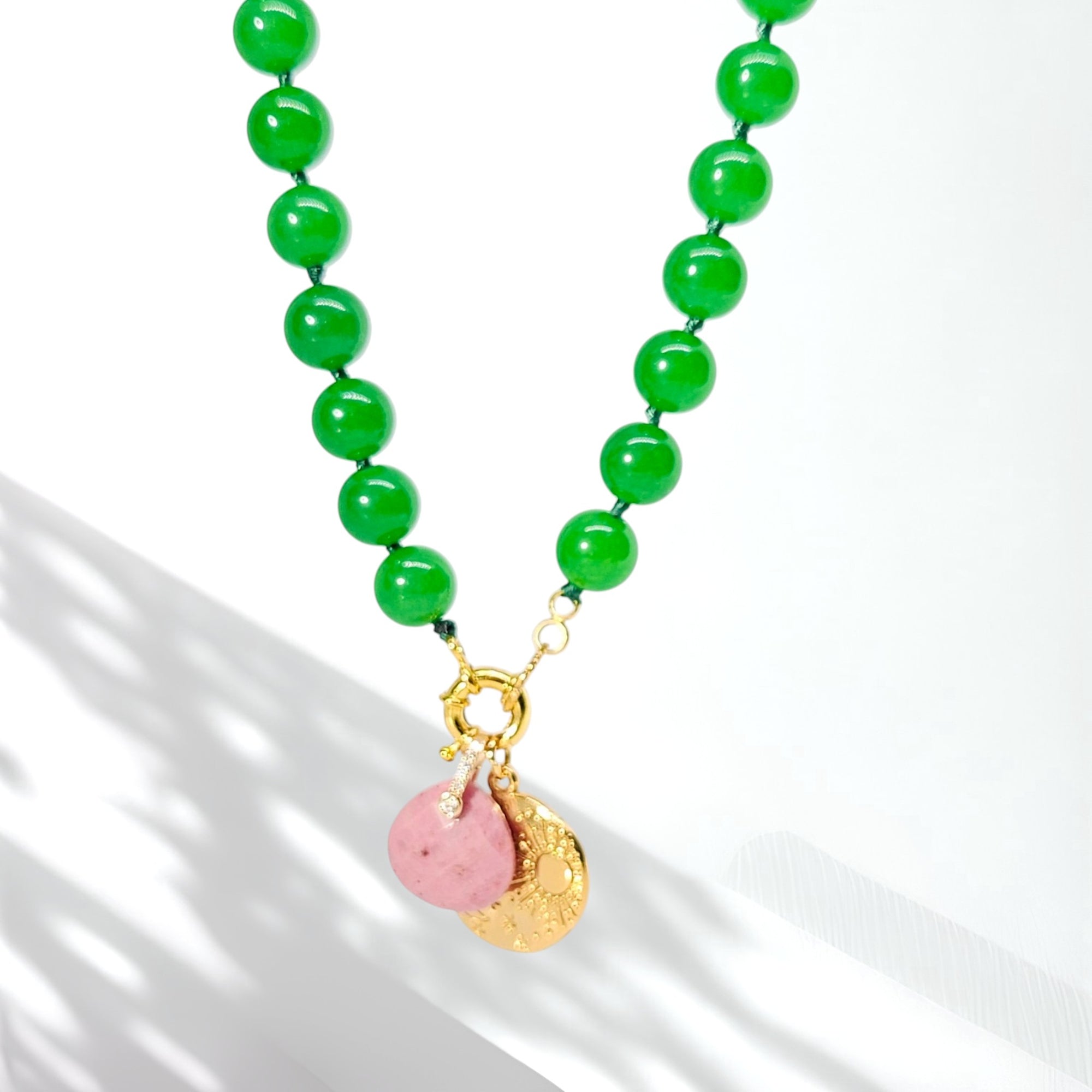 A beaded necklace featuring vibrant green round beads, a gold-tone clasp, and two small hanging charms: one smooth pink stone and one textured gold disc, displayed on a clean white background with soft shadows.