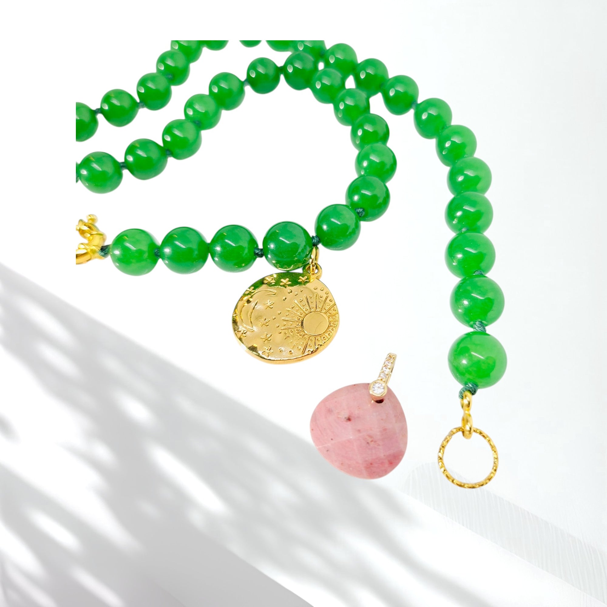 A beaded necklace with vibrant green round beads, gold-toned metal details, a round gold sun pendant, and a pink stone pendant, displayed on a white surface with soft shadows.