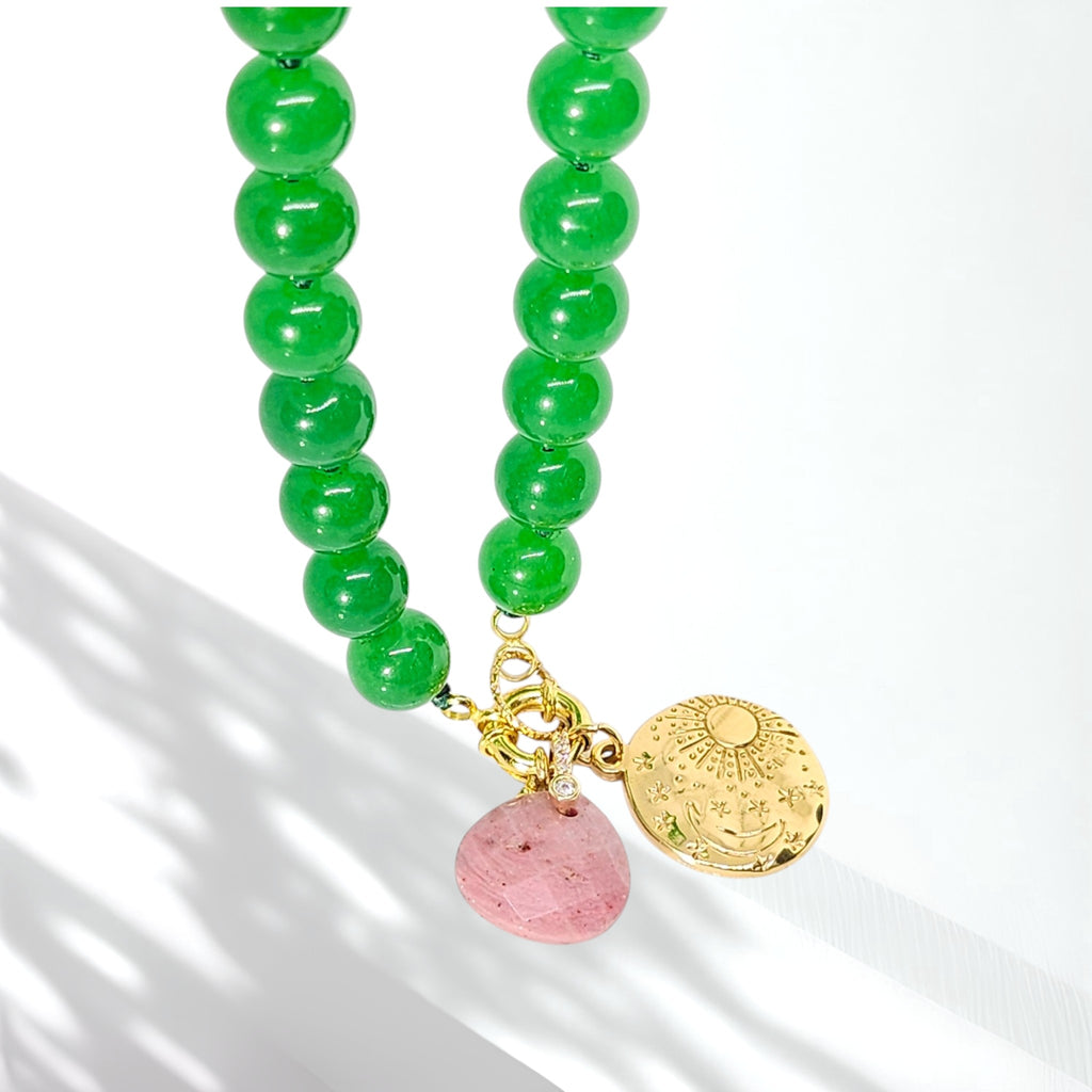 A beaded necklace with glossy green round beads, a small gold-tone charm, a pink faceted stone bead, and a round gold-tone medallion pendant, photographed on a white background with soft shadows.