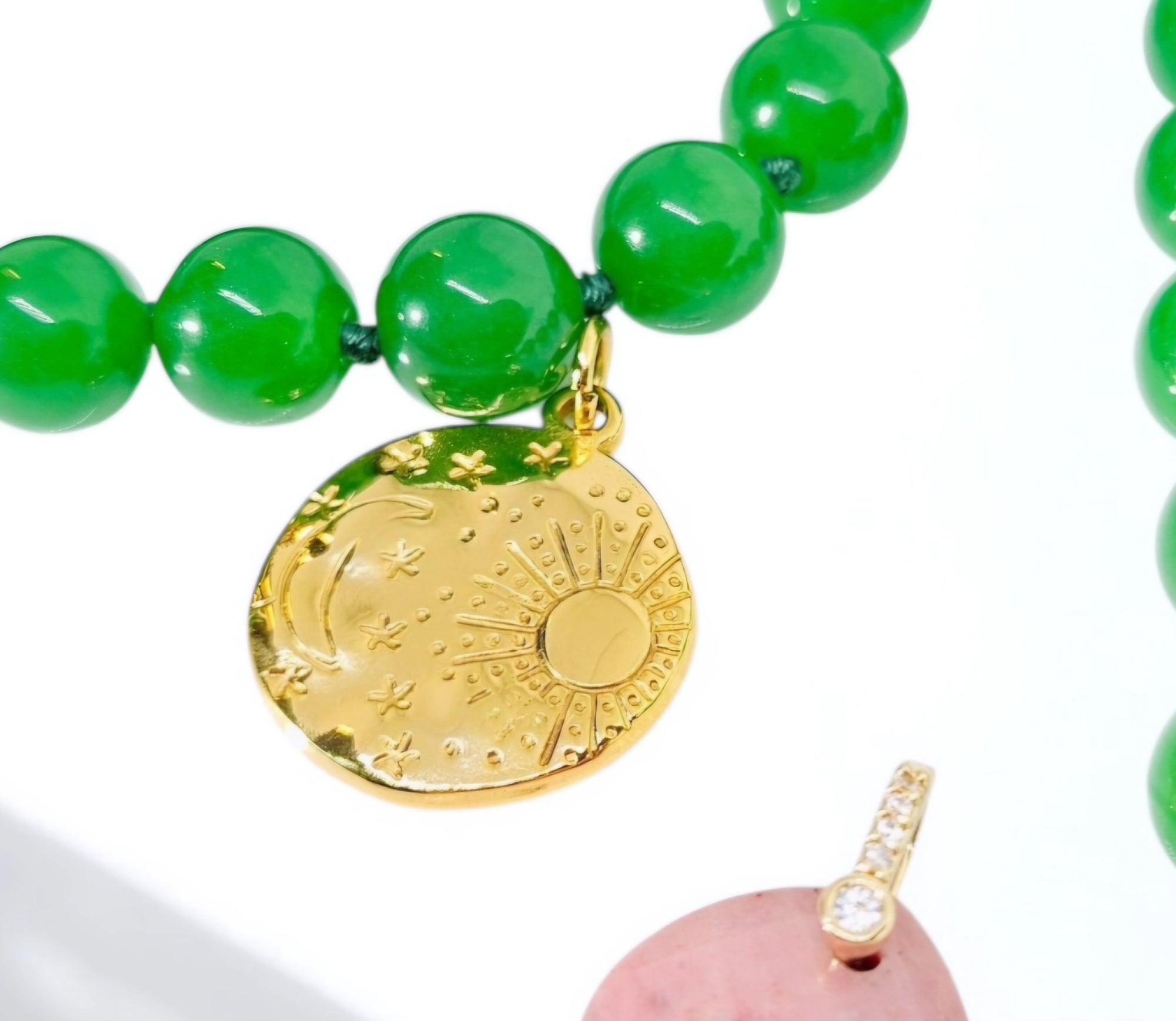 A close-up of a beaded necklace with glossy green round beads and a gold-toned oval pendant engraved with a sun and moon design. A small pink stone pendant with a clear stone clasp is partially visible at the bottom right.
