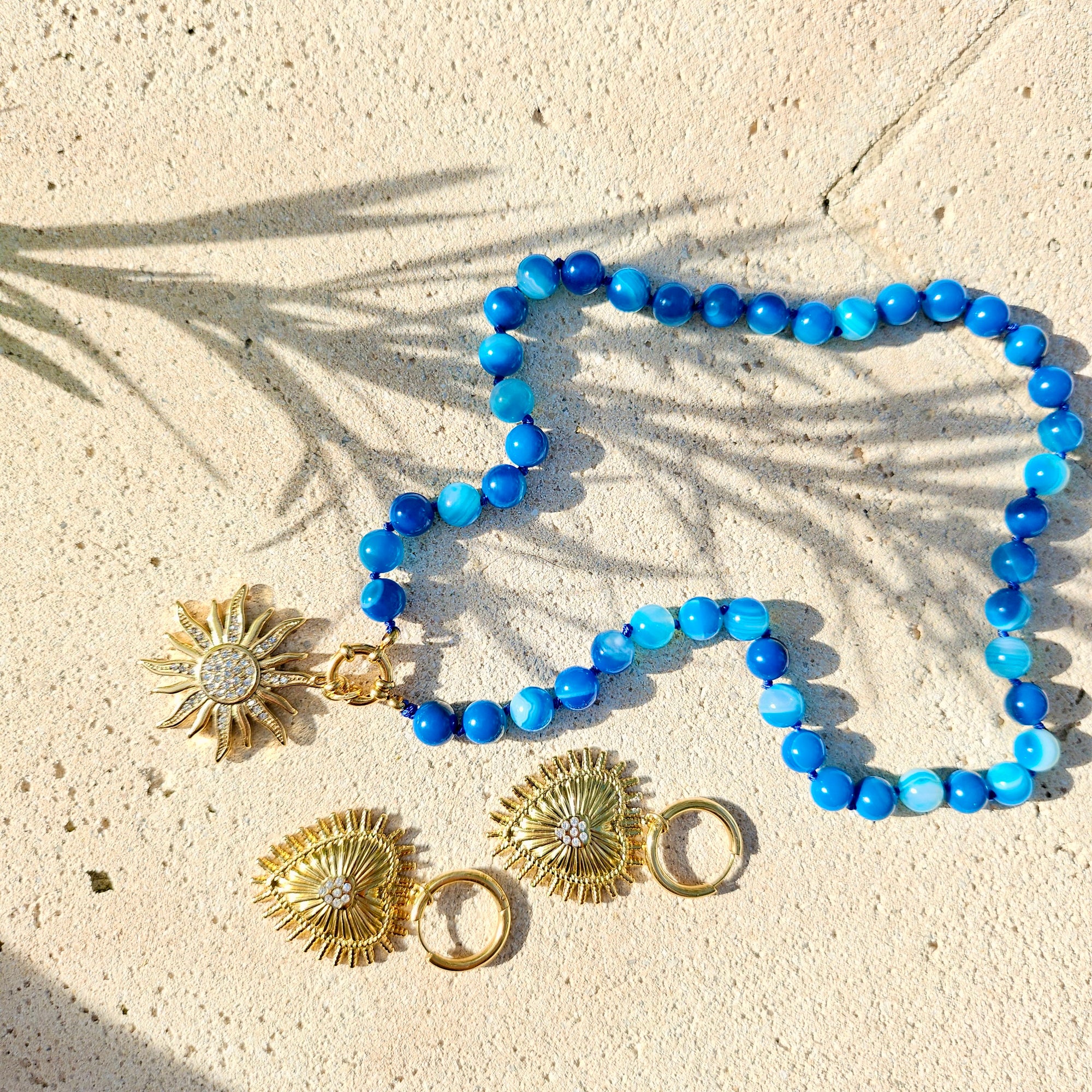 A blue beaded necklace with a gold sunburst pendant and matching gold earrings with clear crystal accents, arranged on a sandy surface with palm leaf shadows.