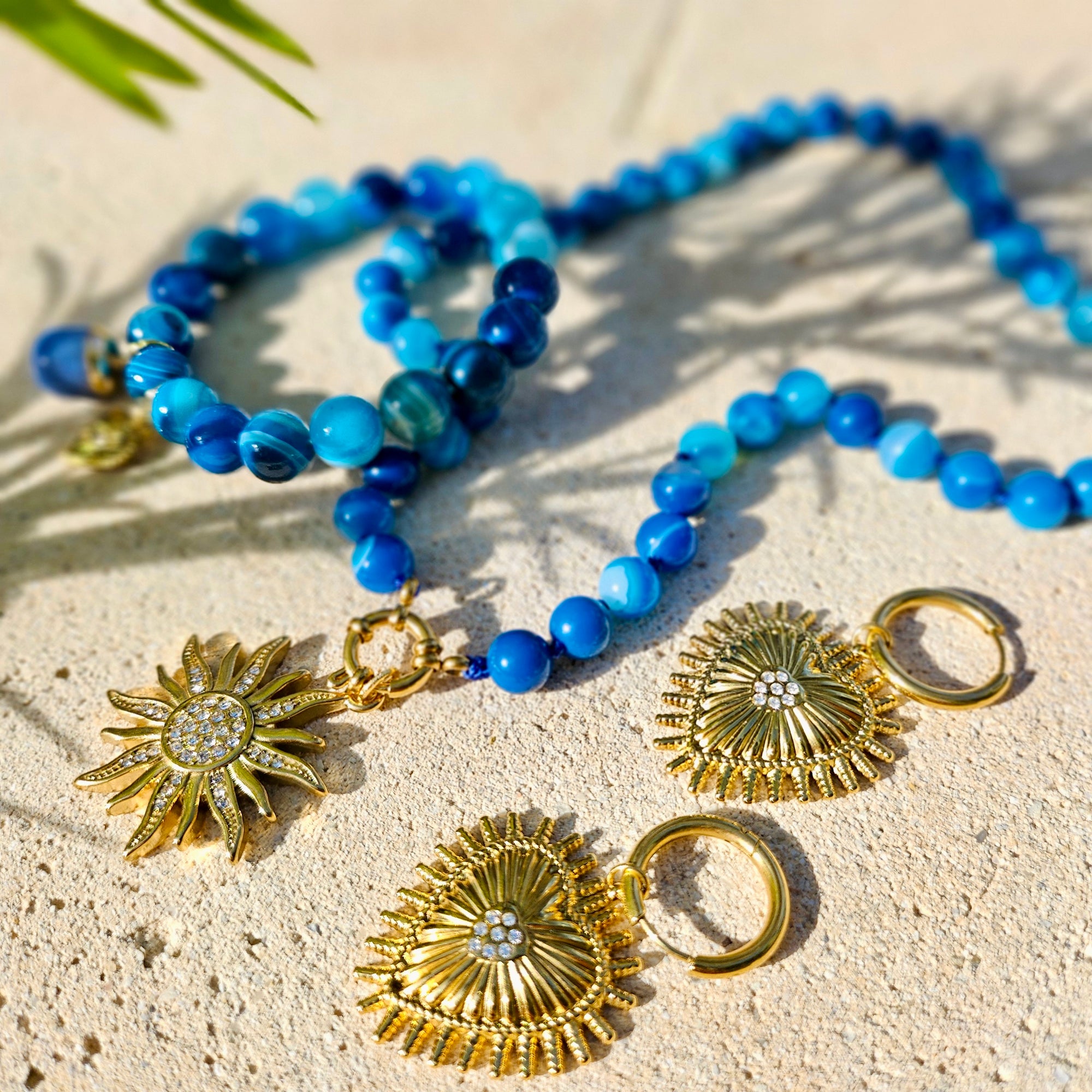 Blue beaded necklace and matching earrings with gold sunburst charms, arranged on a light sandy surface with soft shadows.
