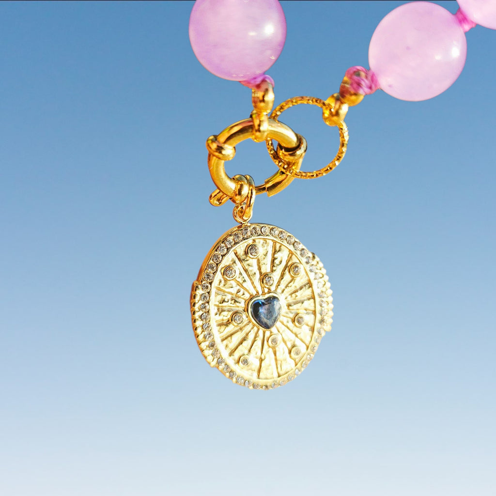 Gold-toned pendant jewelry featuring a circular sunburst medallion with a small dark heart-shaped center, hanging from pink translucent beads and a gold clasp against a blue gradient background.