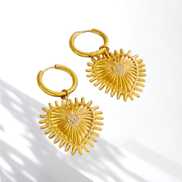 A pair of gold hoop earrings with textured, sunburst-shaped heart pendants featuring a small clear crystal center, displayed on a clean white background with soft shadow overlays.