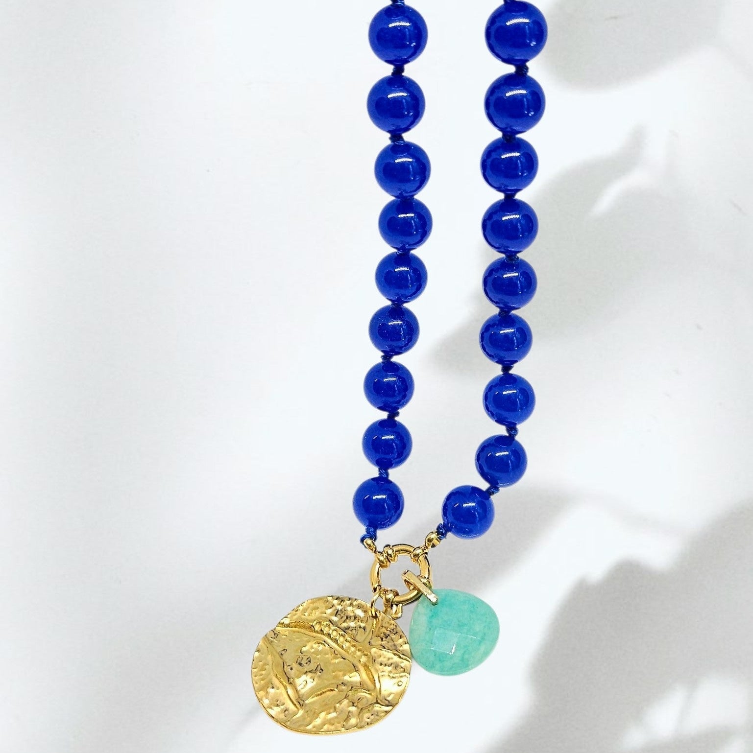 A beaded necklace featuring two strands of polished royal blue beads, a textured gold coin pendant, and a small round light green stone accent, displayed against a white background with soft shadows.