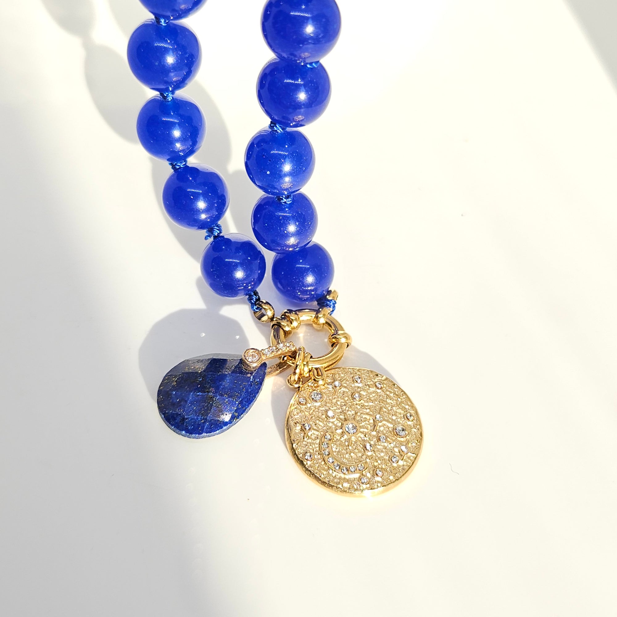 A close-up of a beaded necklace with glossy cobalt blue round beads, a gold-tone clasp, a round textured gold charm, and a faceted dark blue teardrop gemstone on a white background.