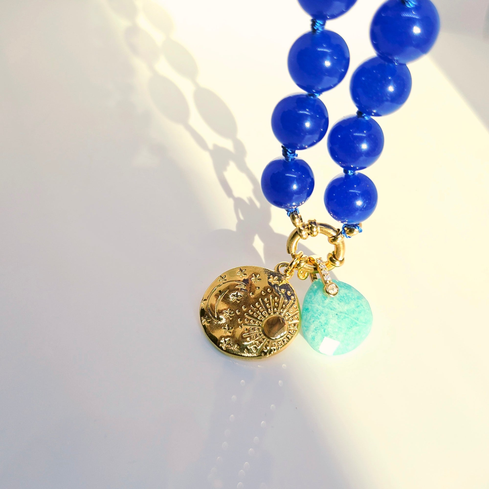 A close-up of a beaded necklace with glossy cobalt blue beads, a gold-tone sunburst charm, and a light green faceted bead, photographed on a bright white surface with strong sunlight and shadows.