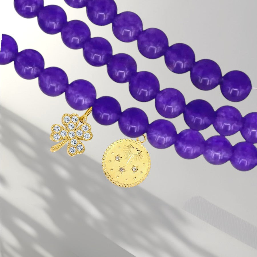 Purple beaded necklace with two gold charms, one clover-shaped and one round medallion, on a light gray background.
