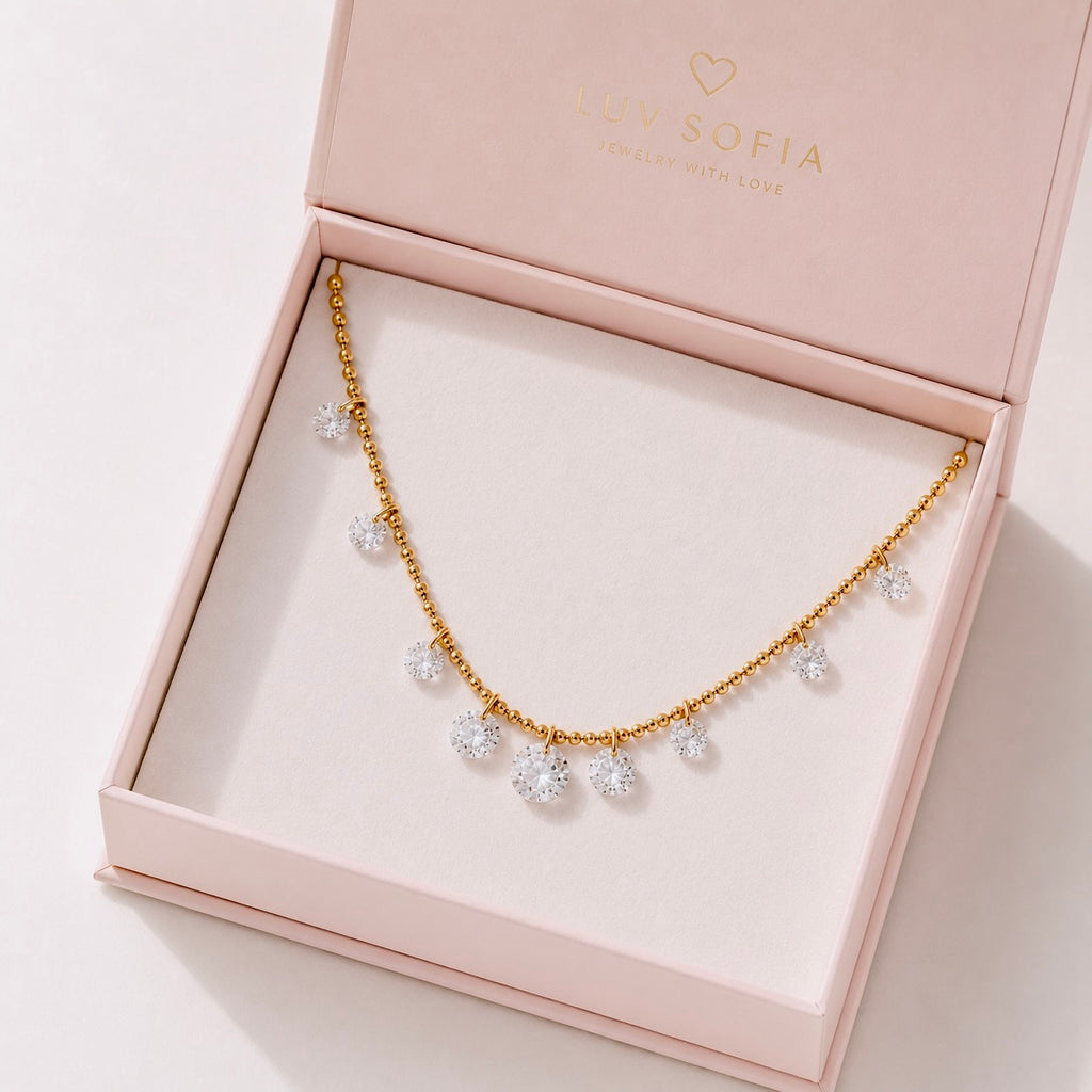 A gold chain necklace with small clear crystal flower-like charms, presented in a light pink Luv Sofia jewelry box on a white background.
