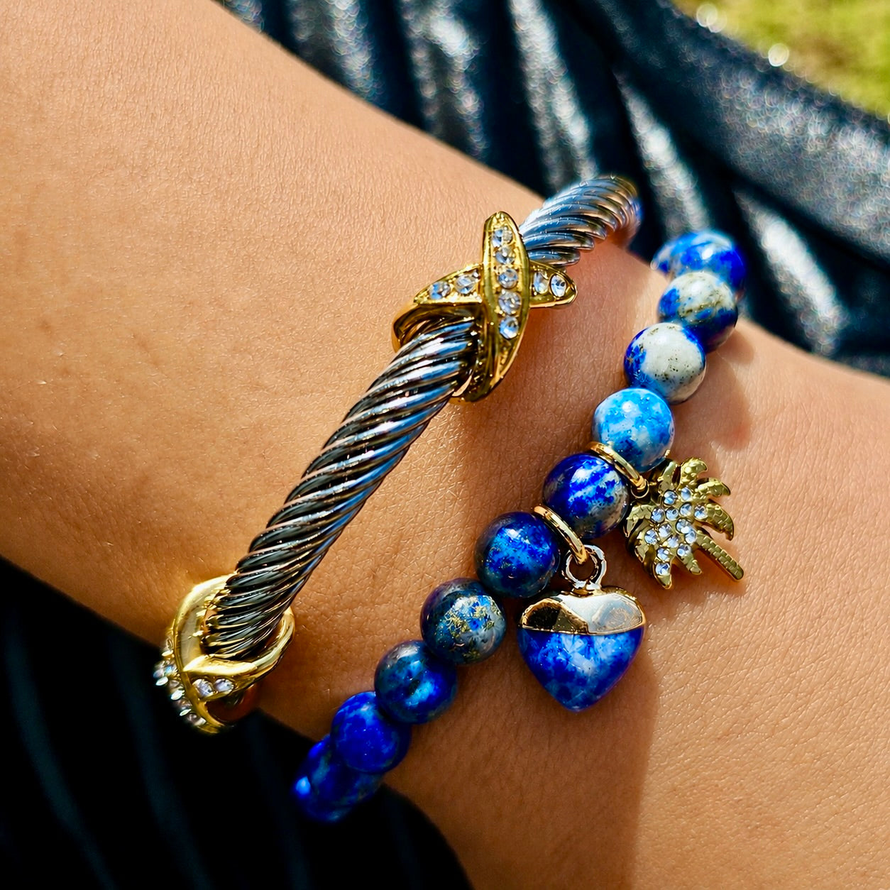 A close-up of a person's wrist wearing two bracelets: one is a silver-toned twisted cable bracelet with gold-toned crystal-embellished end caps and a cross charm, and the other is a beaded bracelet with blue and white marbled beads, a gold palm tree charm, and a gold heart charm.