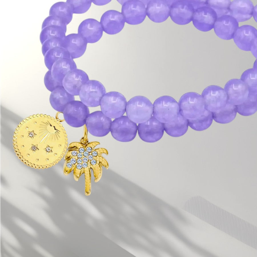 A purple beaded stretch bracelet with two gold charms, one a round sunburst medallion and the other a palm tree adorned with small clear crystals, shown on a light gray background.