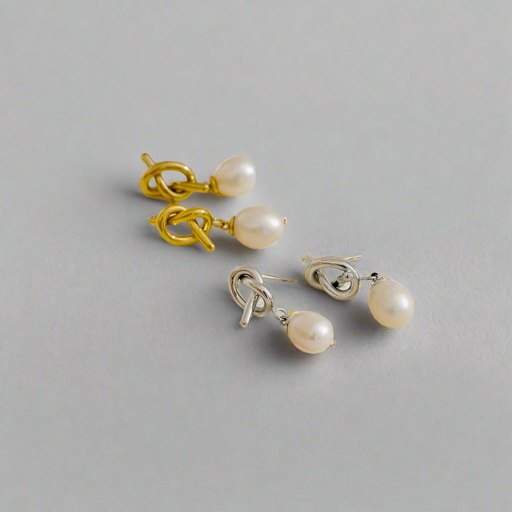 two pairs of drop earrings featuring genuine freshwater pearls suspended from a polished knot detail, on a light grey background. 