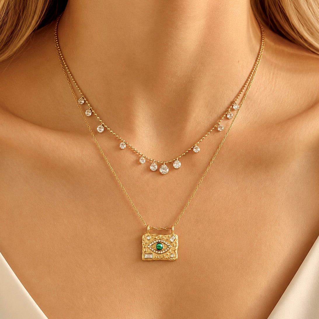 A close-up of a model wearing a delicate gold double-layer necklace. The top layer features small clear crystal drops, while the bottom layer has a textured gold rectangular pendant with a green evil eye design surrounded by tiny clear crystals.