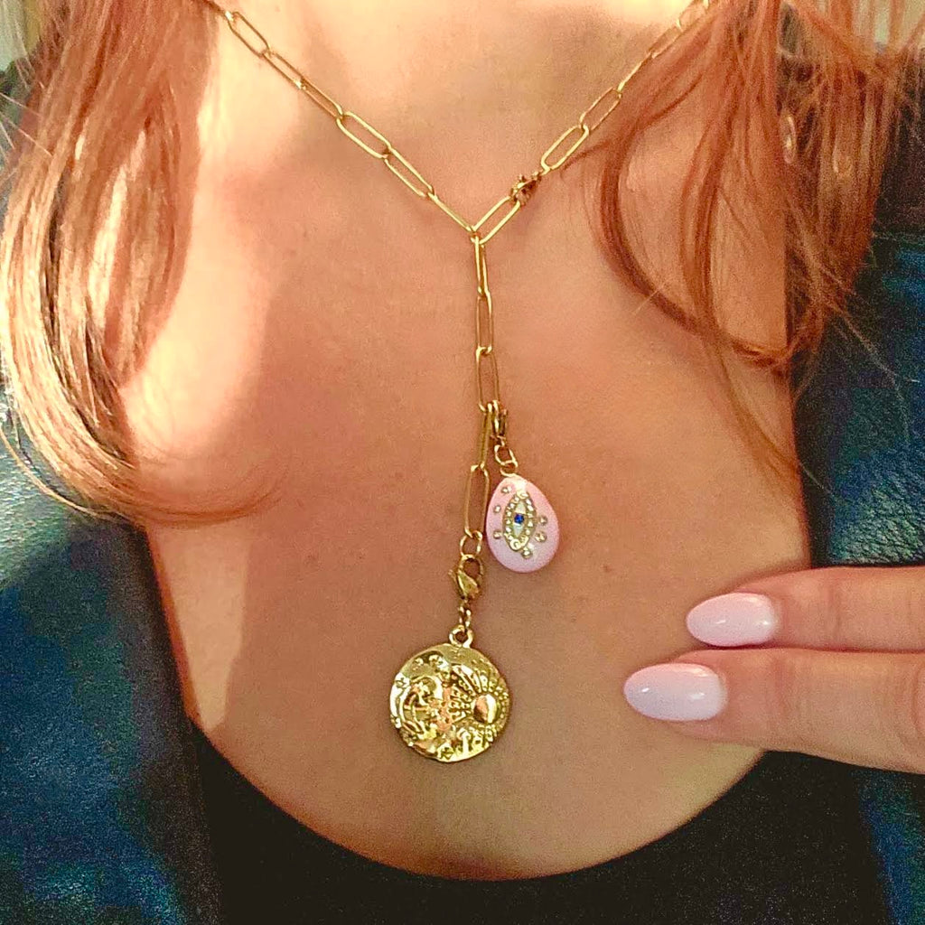 A close-up of a gold chain necklace worn on a person's neck, featuring a round gold pendant and a small pink teardrop charm with a decorative center.