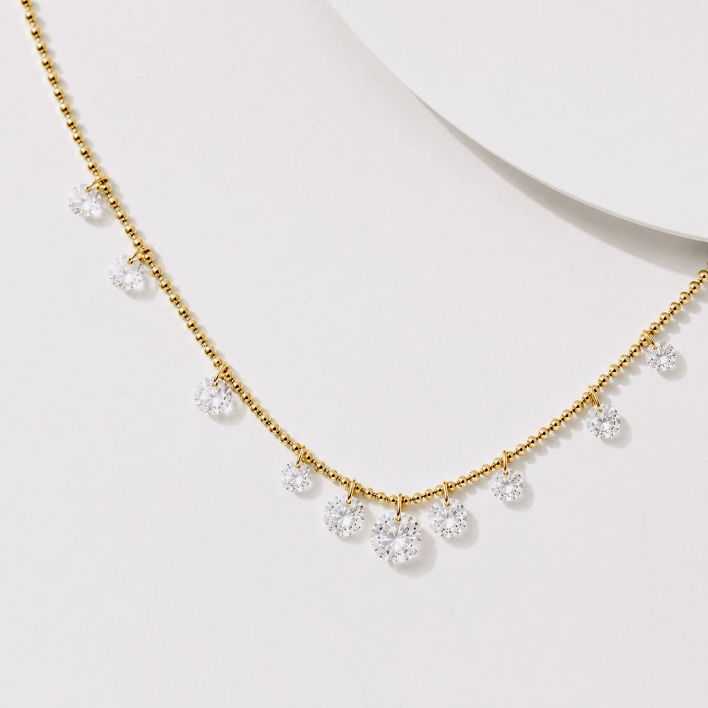A delicate gold necklace with small clear gemstone clusters hanging along the front, displayed on a clean white surface.