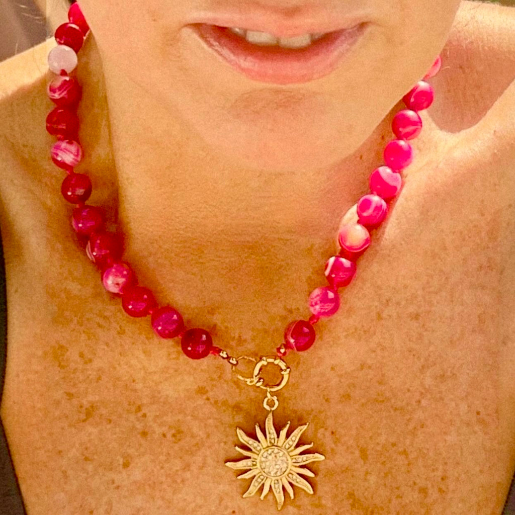 A close-up of a person wearing a beaded necklace with bright pink and magenta round beads, a gold clasp, and a gold sunburst pendant with a clear central stone.