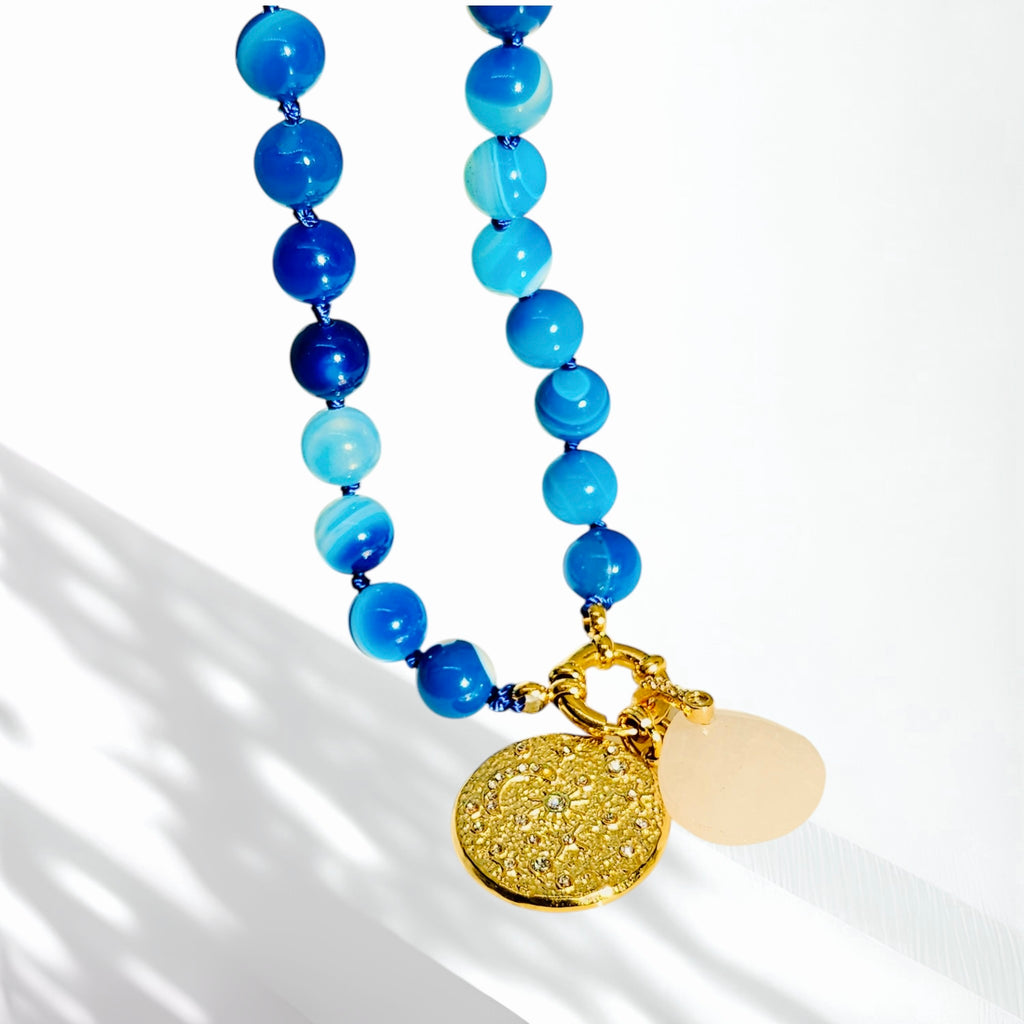 A beaded necklace with blue and light blue round beads, a gold-tone clasp, a textured gold disc pendant, and a pale pink stone accent, photographed on a white background with soft shadows.