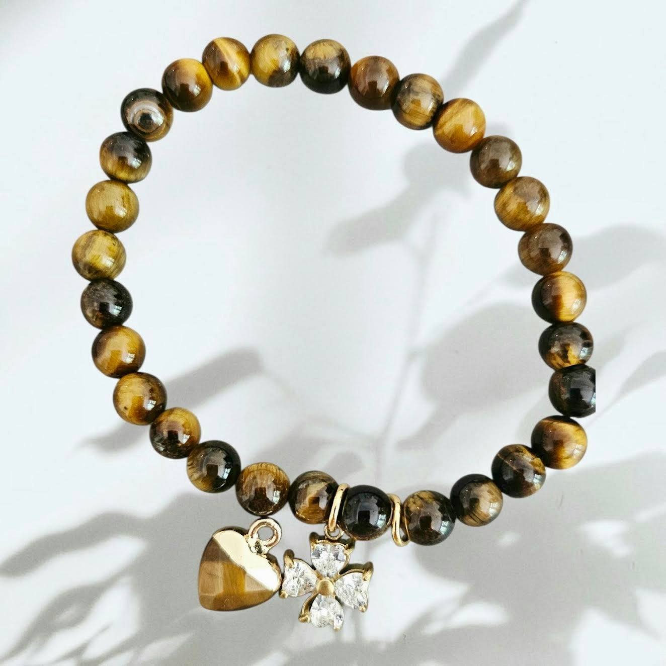 A beaded stretch bracelet made of polished tiger's eye beads in brown and black, featuring a gold heart charm and a gold flower charm with a clear rhinestone, displayed on a white background with soft leaf shadows.