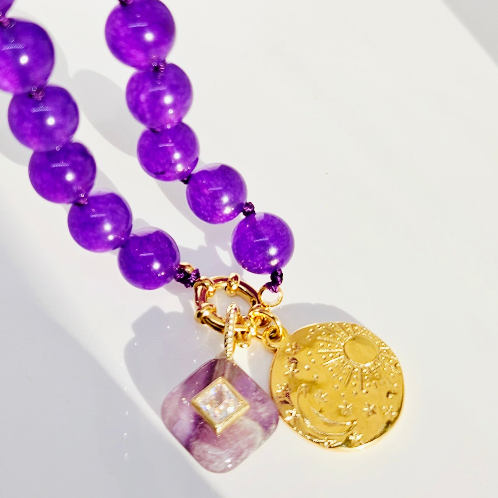 Purple beaded necklace with a gold clasp, featuring a round gold moon and sun pendant and a square purple crystal charm with a clear center stone, shown on a white background.